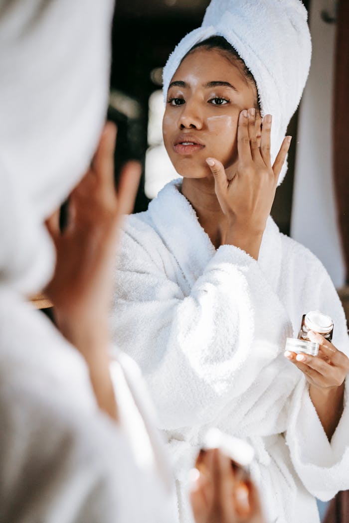 about-us-02 Young black female  in white robe and towel on head  applying moisturizing cream on face while standing in bathroom